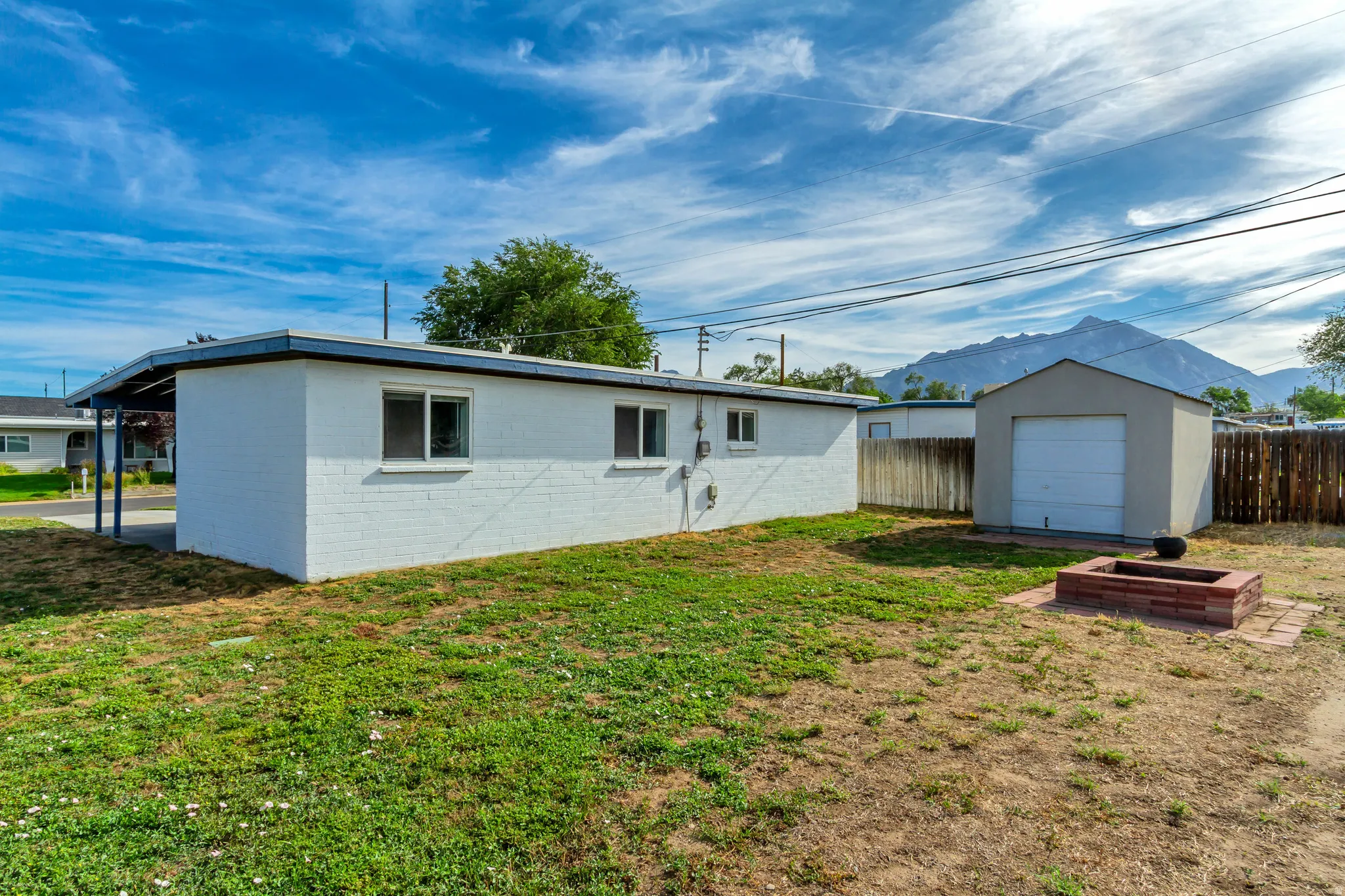 Rear exterior featuring white painted brick, a covered carport, a detached shed, and a brick fire pit