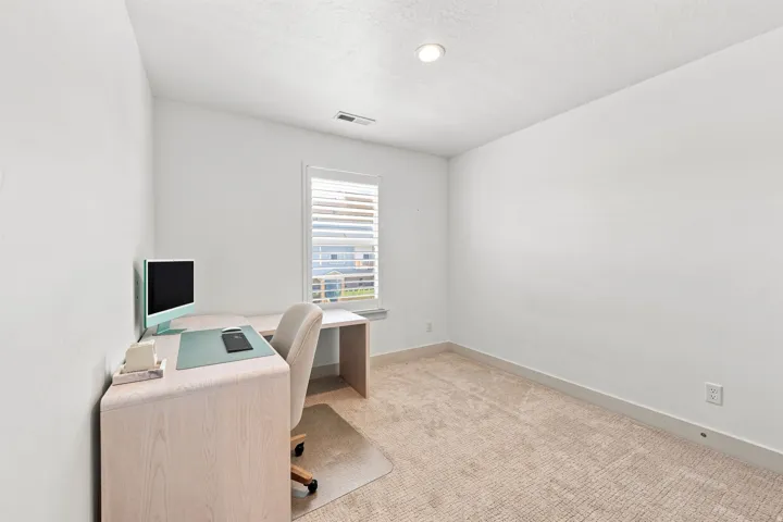 Bright home office featuring neutral carpeting, light wood-finish desk with integrated workspace, and a large window with plantation shutters providing natural illumination