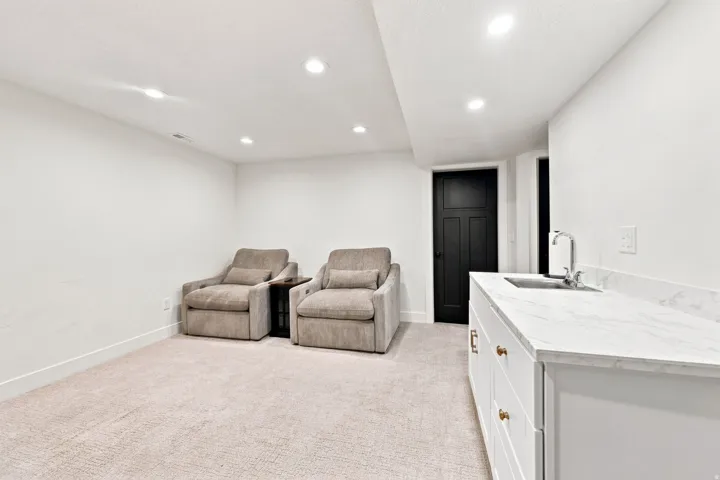 Sophisticated recreation space featuring recessed ceiling lighting, neutral carpet flooring, and a custom white cabinetry wet bar with a polished countertop and integrated sink