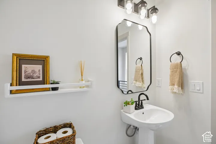 Pedestal sink with a black-finish faucet, framed mirror, wall-mounted light fixture, and a floating shelf