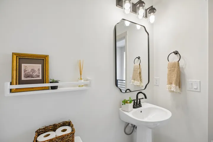 Pedestal sink with a black-finish faucet, framed mirror, wall-mounted light fixture, and a floating shelf