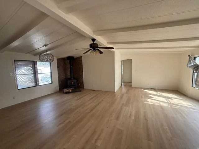Vaulted ceilings with exposed beams, wood-finish flooring, wood-burning stove with stone-finish surround, and a ceiling fan