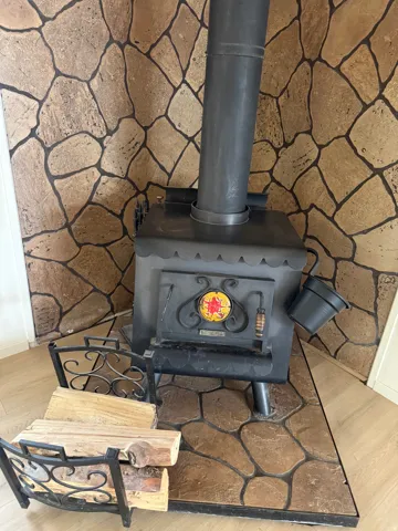 Black cast iron wood-burning stove with a decorative door, set against a stone-patterned wall, and grounded by a stone-patterned hearth