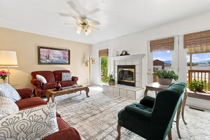 Warm living space featuring a tiled fireplace with a white mantel, ceiling fan, and two glass-paneled doors with natural woven blinds