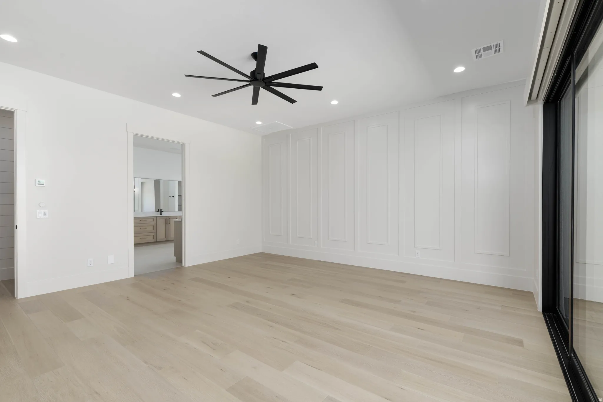 Light wood-finish flooring, accent wall with decorative molding, recessed ceiling lighting, ceiling fan with matte black finish, and large sliding glass door
