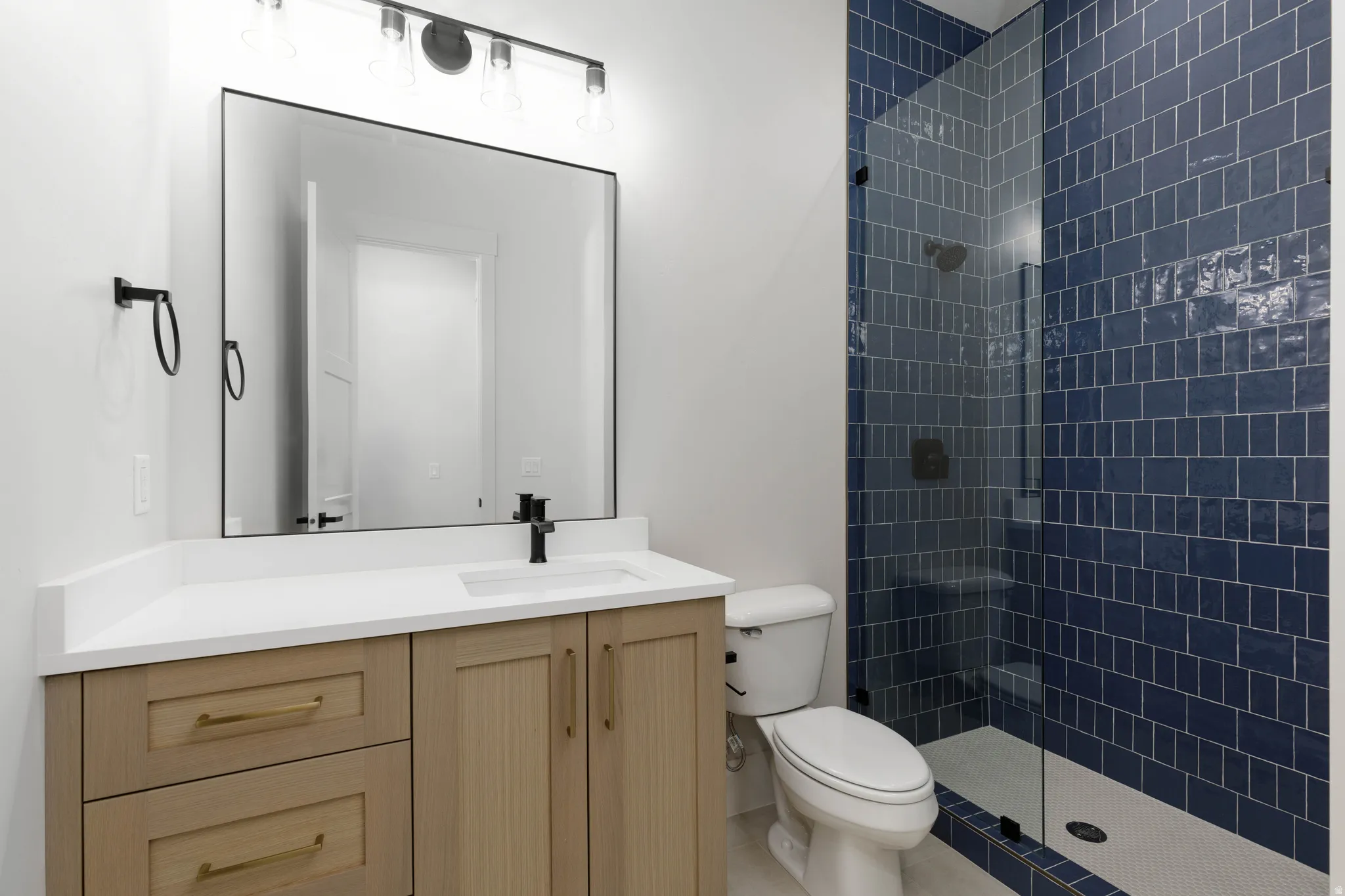 Vanity with light-toned cabinetry and white countertop, black fixtures, frameless mirror, and a walk-in shower with blue subway tile