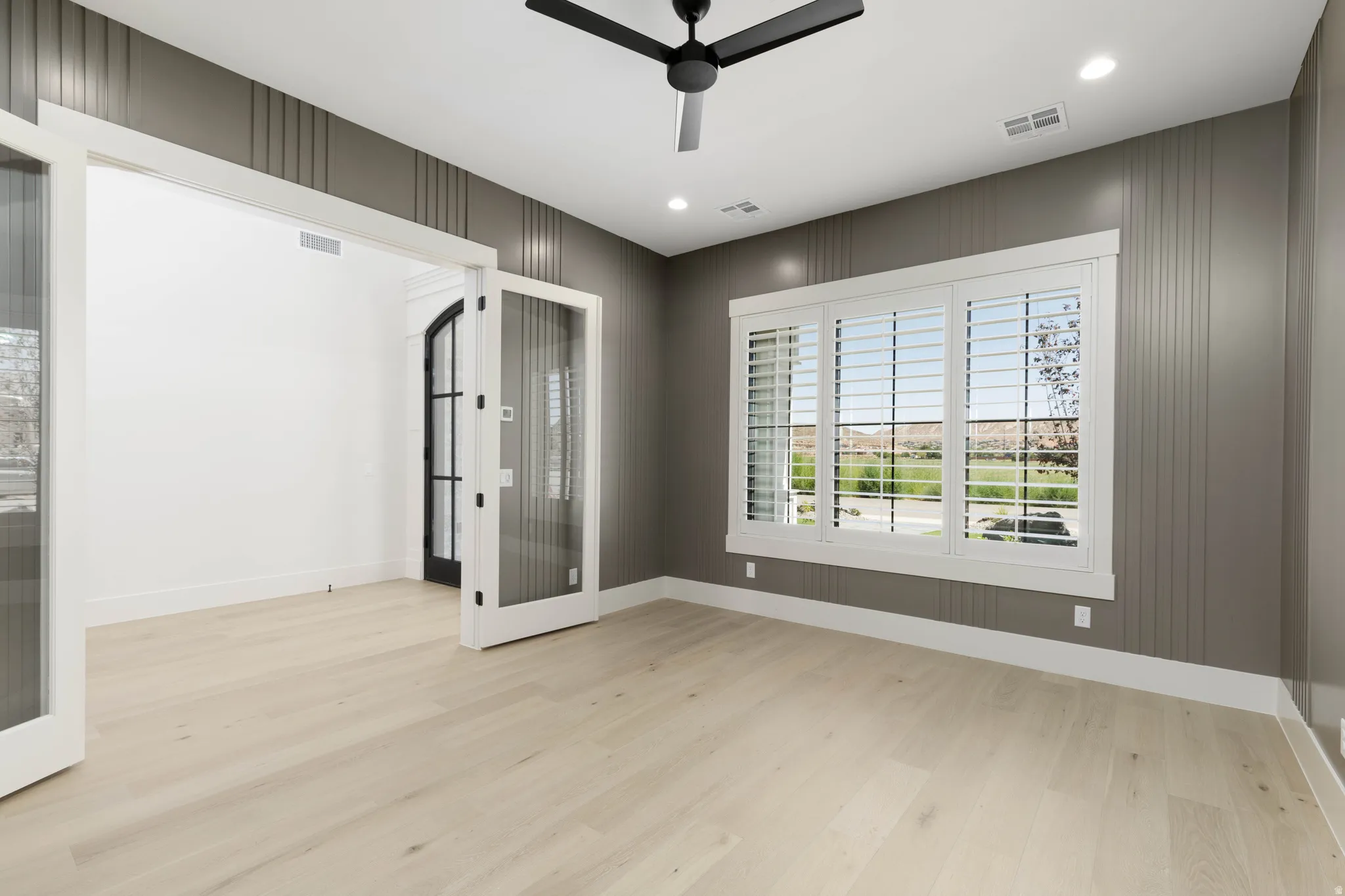 Light wood-finish flooring, accent wall with vertical paneling, large window with white plantation shutters, white framed French doors, and a black ceiling fan