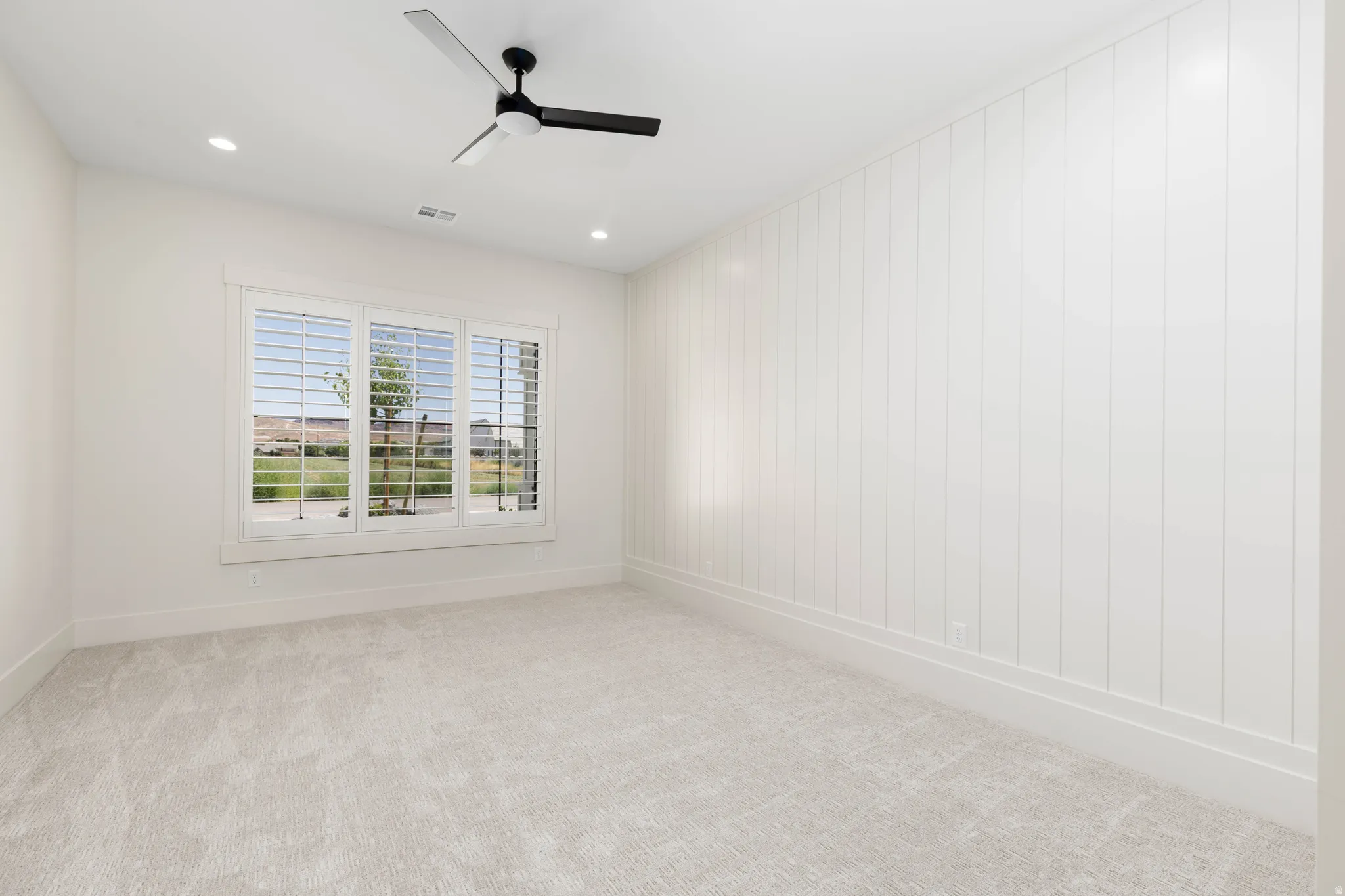 Carpeted room featuring a white paneled accent wall, a three-panel window with plantation shutters, recessed lighting, and a ceiling fan