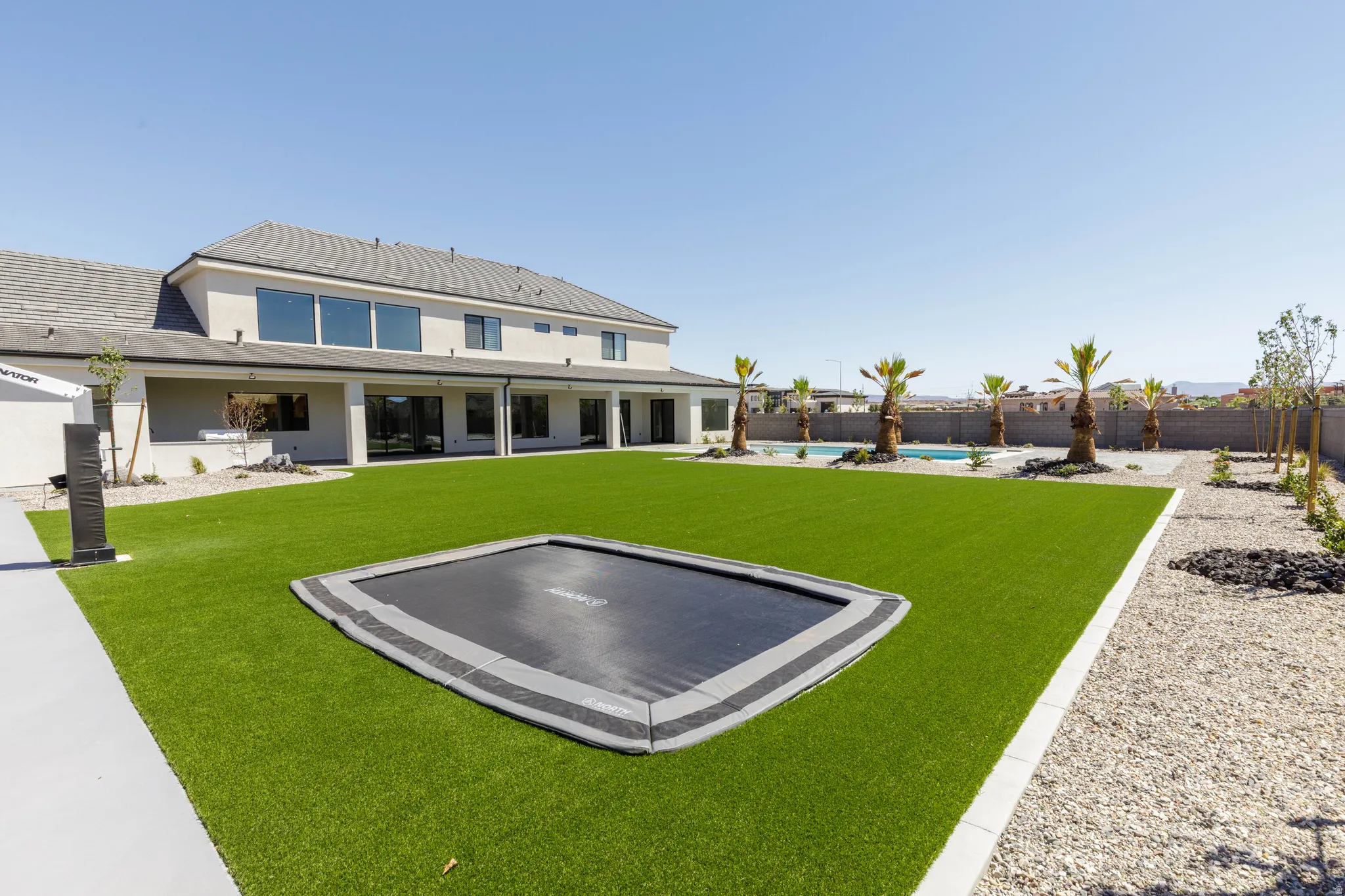 Expansive backyard with artificial turf, built-in trampoline, gravel landscaping, and a swimming pool bordered by palm trees