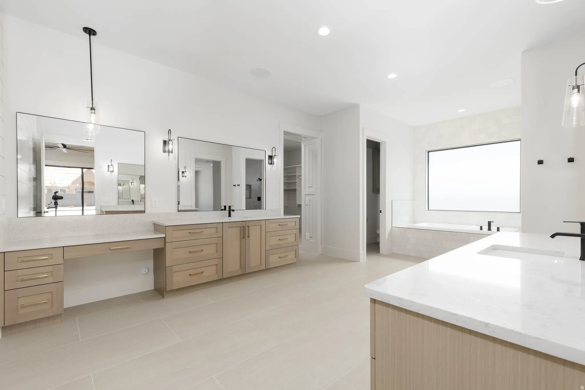 Light-toned flooring, extensive dual vanity with light wood-finish cabinetry, built-in makeup vanity, large soaking tub, and a window for natural light