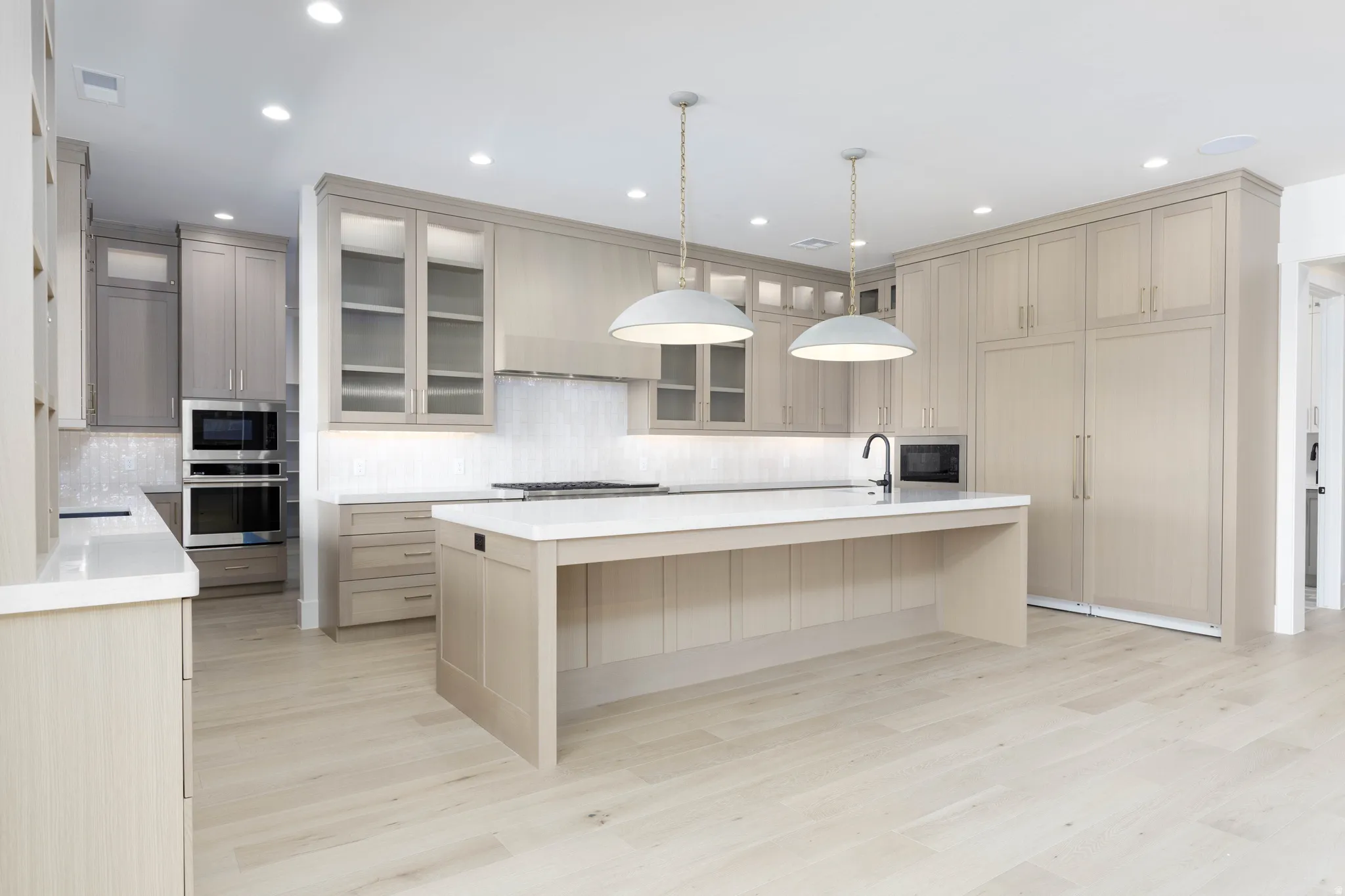 Expansive kitchen featuring light wood-finish flooring, a generous central island with a sink, pendant lighting, built-in double ovens, and extensive cabinetry with integrated appliances