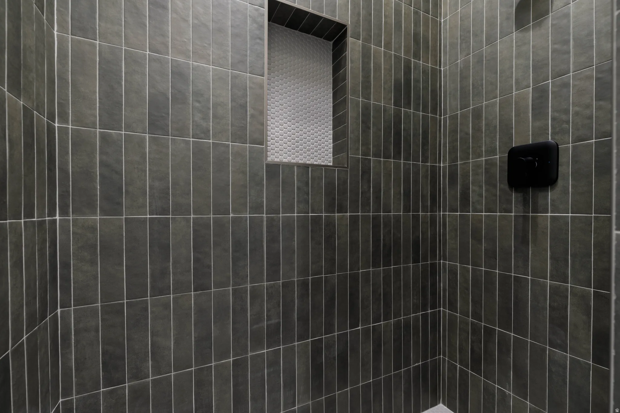 Shower enclosure featuring vertical-stacked gray rectangular tiles, a tiled niche with contrasting mosaic tile, and a matte black shower control fixture