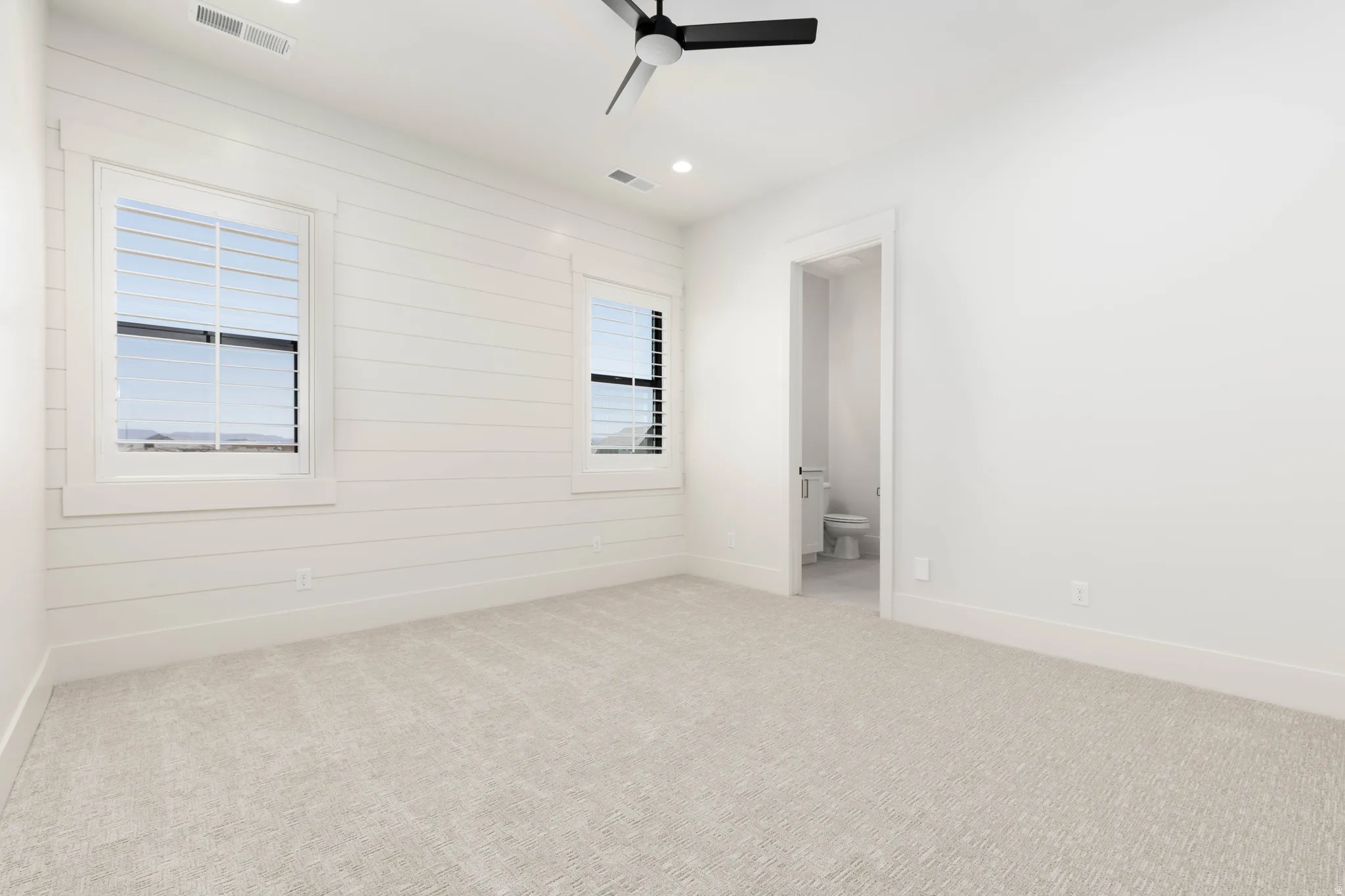 White shiplap accent wall, two windows with plantation shutters, recessed lighting, ceiling fan, and carpeted flooring
