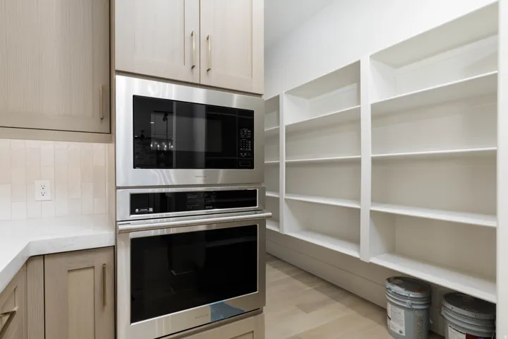 Built-in stainless steel microwave and oven, light wood-finish cabinetry, white solid surface countertop, white subway tile backsplash, and extensive built-in white shelving