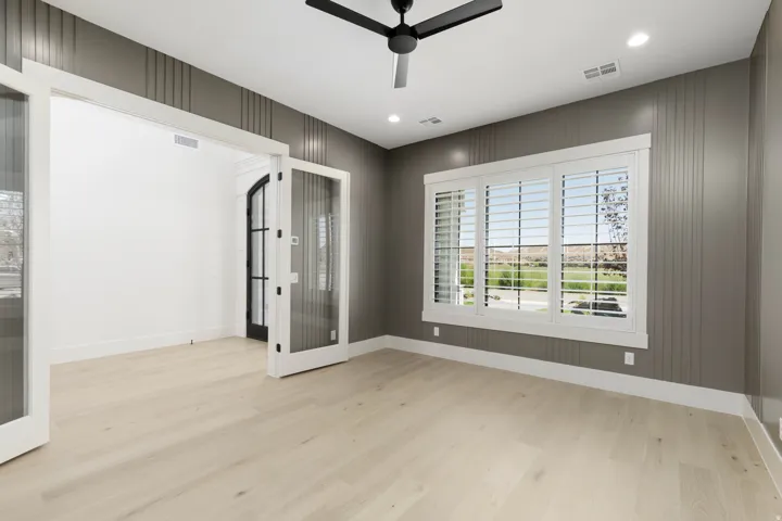 Light wood-finish flooring, accent wall with vertical paneling, large window with white plantation shutters, white framed French doors, and a black ceiling fan