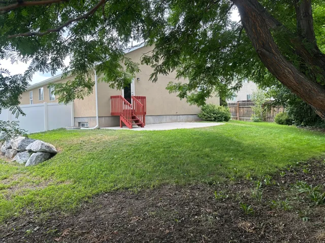 Expansive backyard featuring a manicured lawn, concrete patio, red wood-finish deck, and white privacy fencing