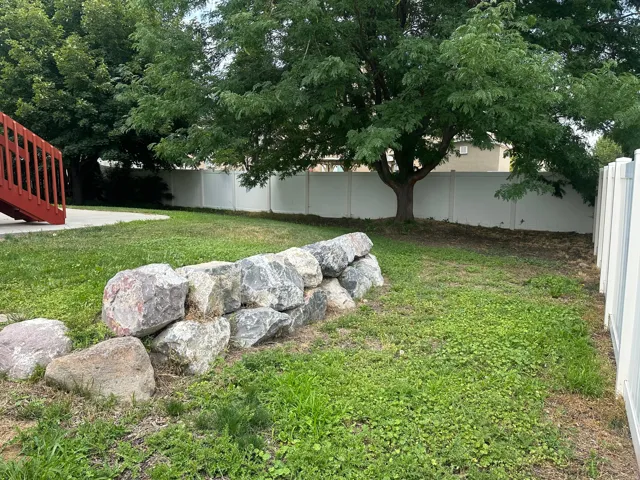 Expansive yard featuring a natural rock retaining wall, mature trees, concrete patio, and privacy fencing