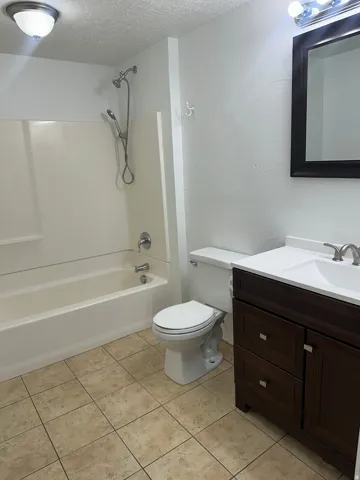 Bathroom featuring a white bathtub with an integrated shower, a dark wood-finish vanity with a white countertop and sink, a toilet, and light-toned square tile flooring