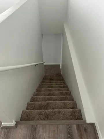 Staircase with carpeted treads, wood-finish flooring, white painted walls, and a wall-mounted handrail