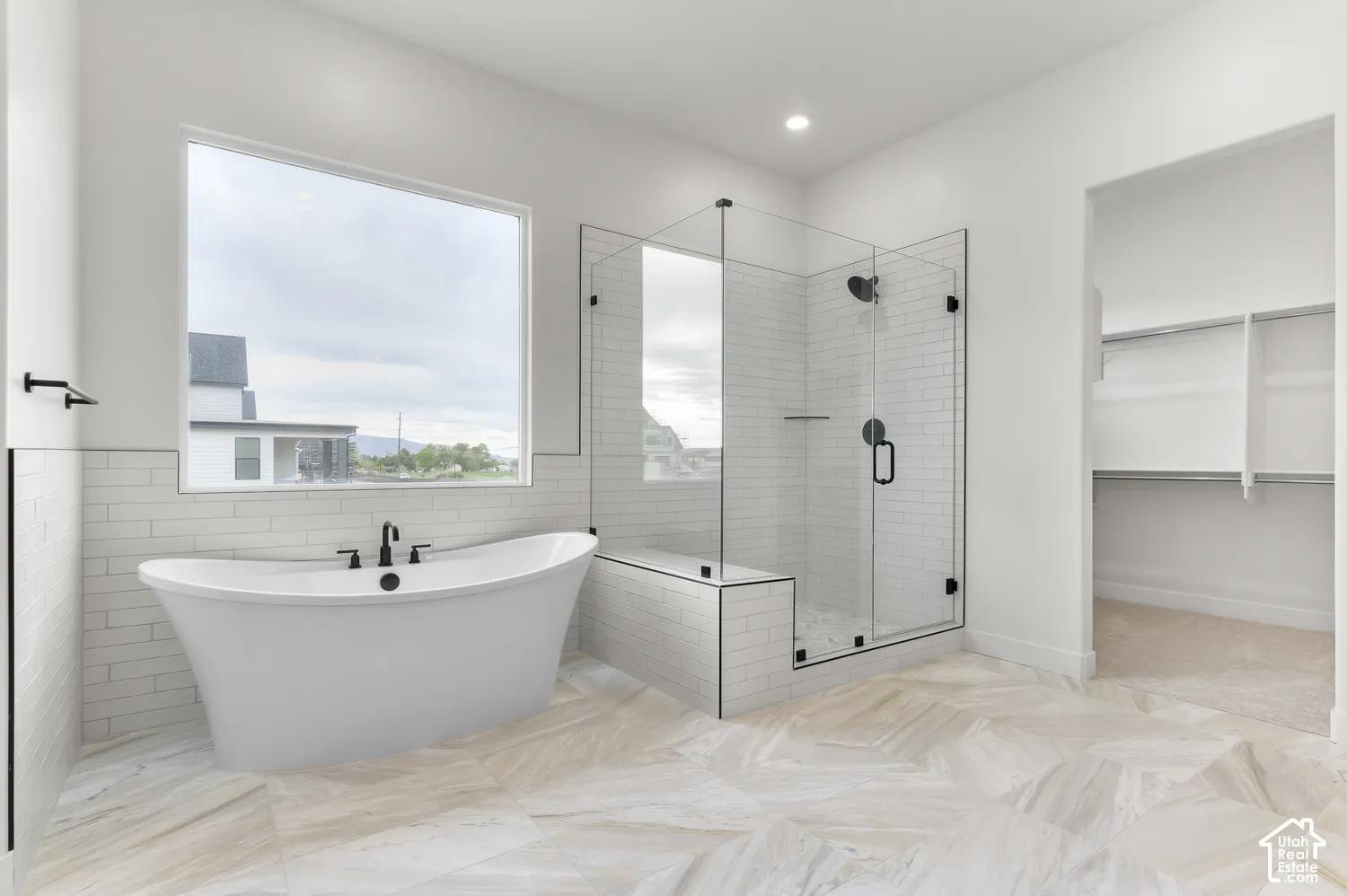 Freestanding soaking tub with matte black fixtures, glass-enclosed shower with subway tile, large window, and built-in closet shelving