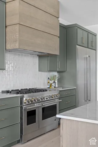 Stainless steel professional-grade range and refrigerator, wood-finish range hood, green cabinetry, and light-toned backsplash