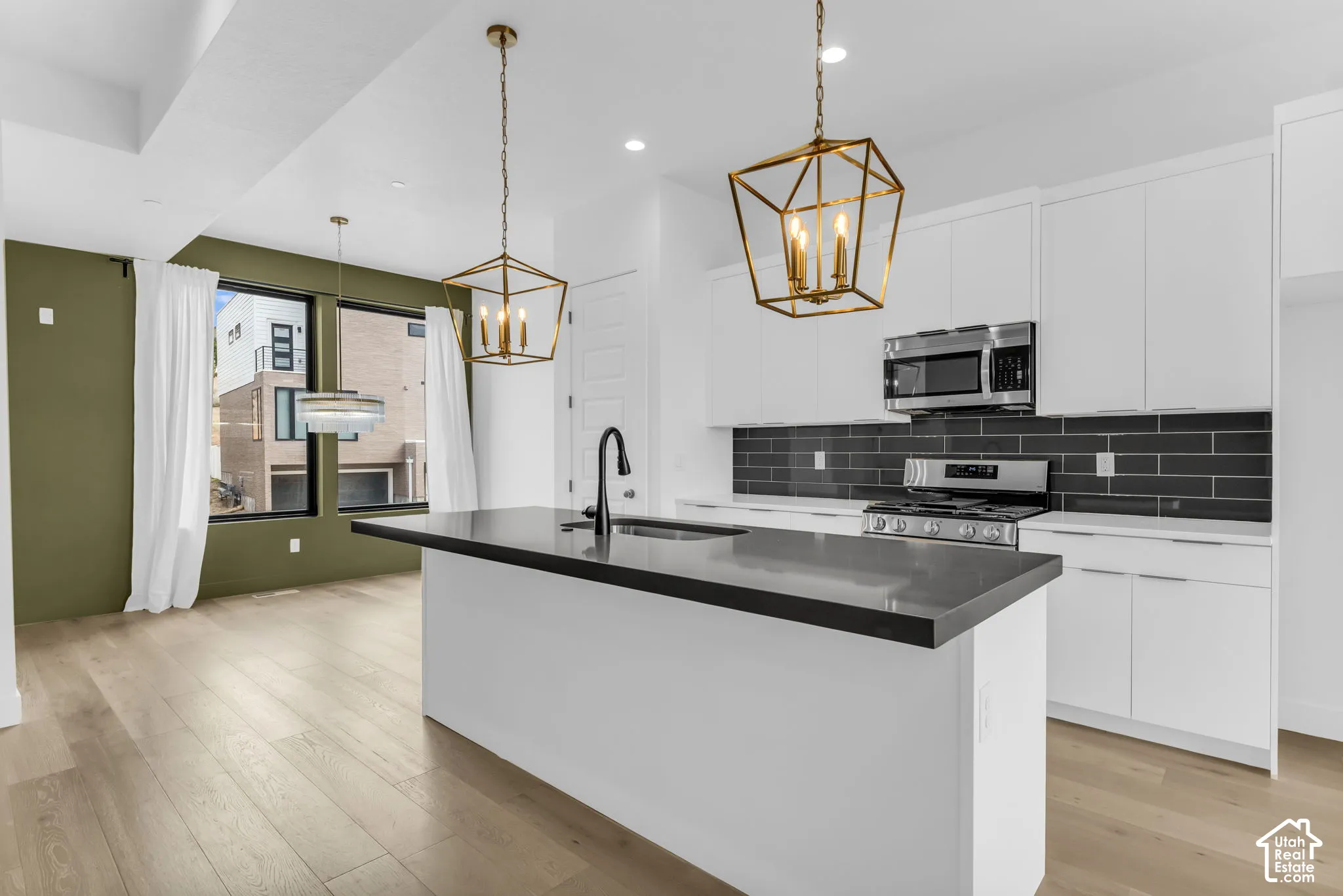 Contemporary kitchen featuring white cabinetry, a waterfall-edge island with black countertops, stainless steel appliances, black subway tile backsplash, pendant lighting with brass accents, and wood-finish flooring throughout the open-concept space