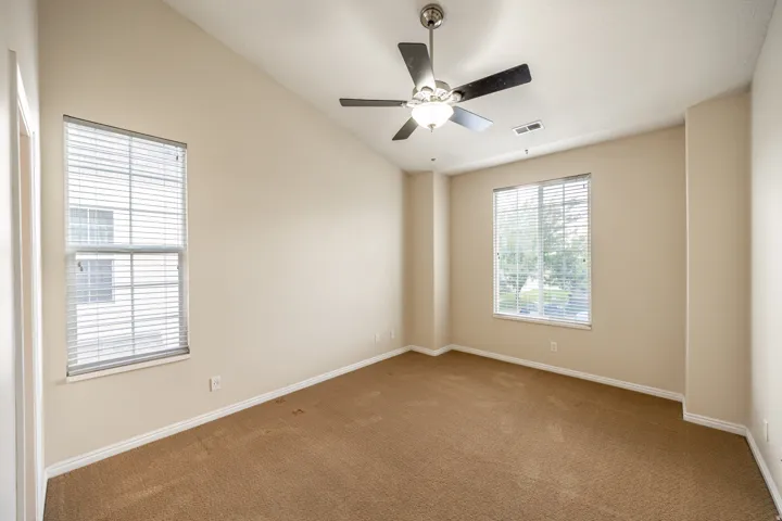 Angled ceiling, two windows with blinds, ceiling fan with light fixture, and neutral carpeting