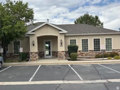 Stucco exterior with stone accent detailing, covered entry, multiple large windows, mature landscaping, and an asphalt parking lot