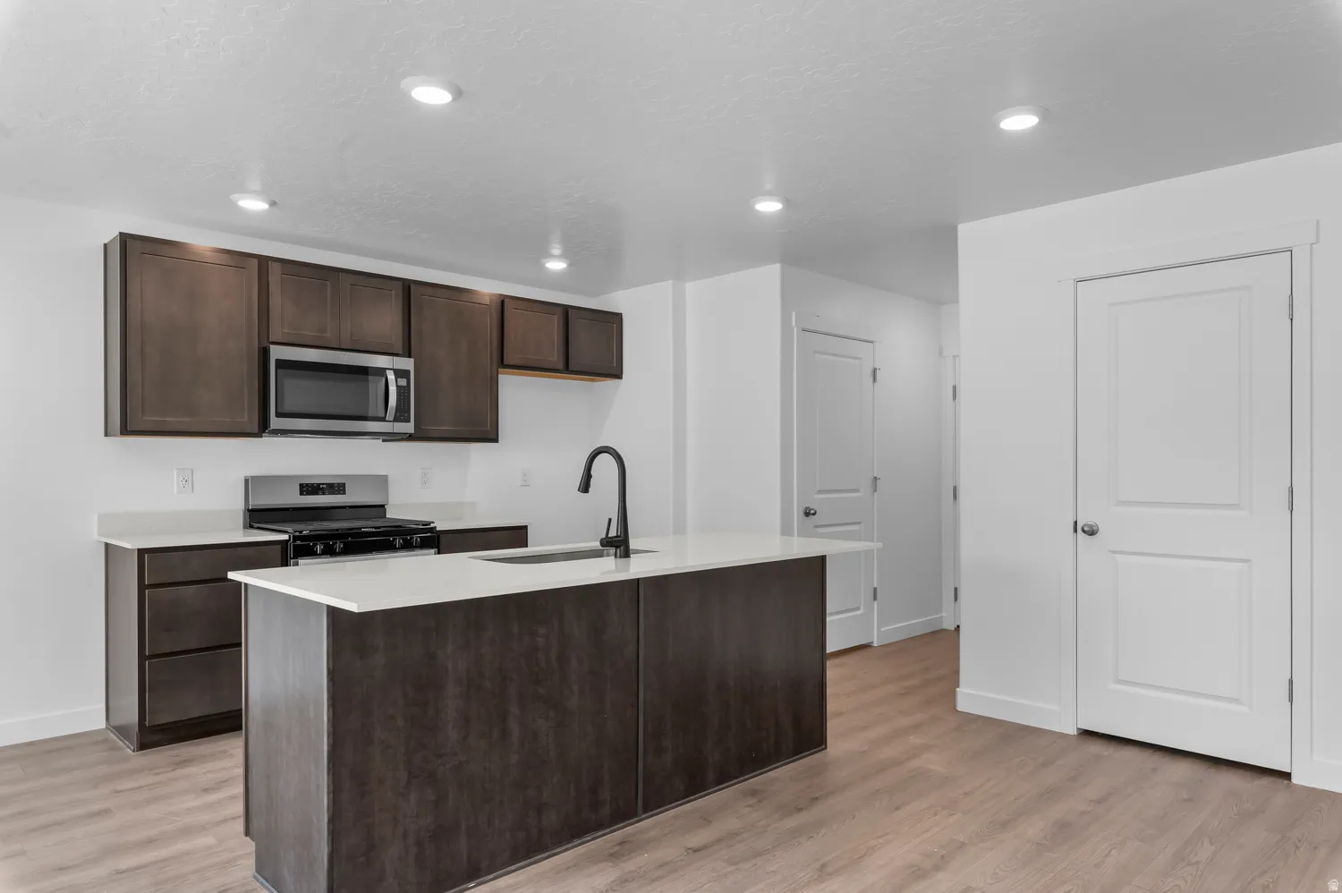 Kitchen featuring dark wood-finish cabinetry, light countertops, stainless steel appliances, a center island with sink, and wood-finish flooring