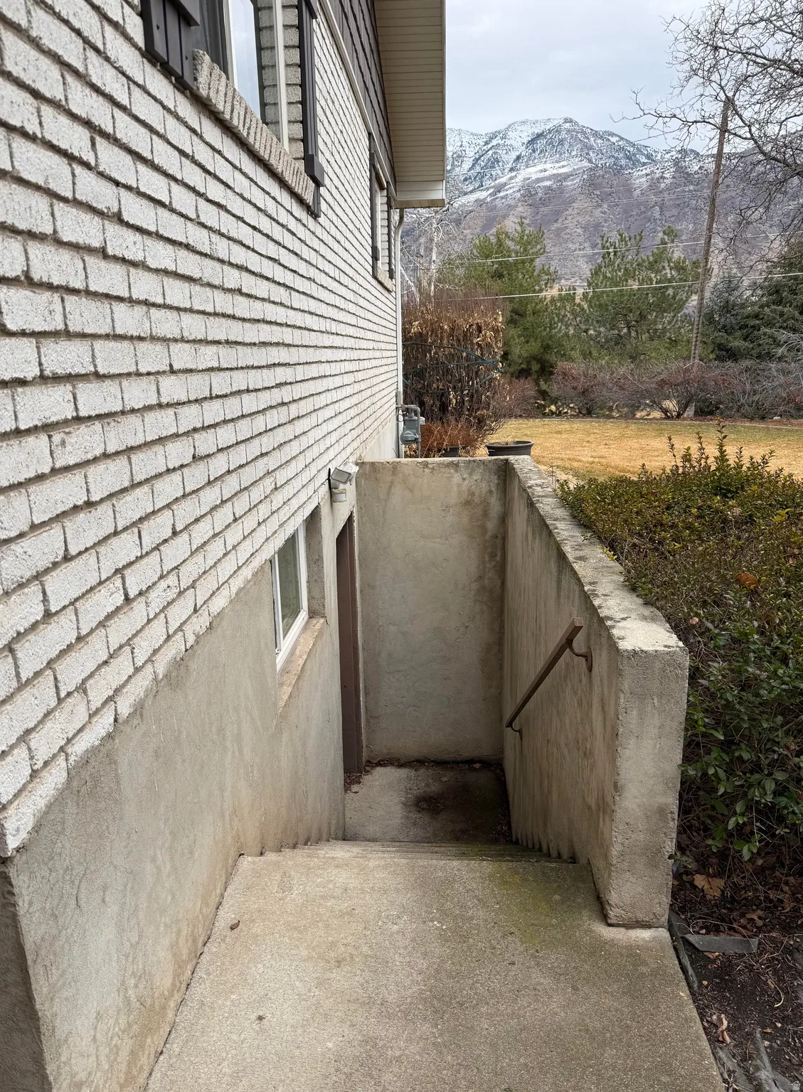 White brick exterior with a concrete foundation, a concrete stairwell with a metal handrail, and a snowy mountain backdrop