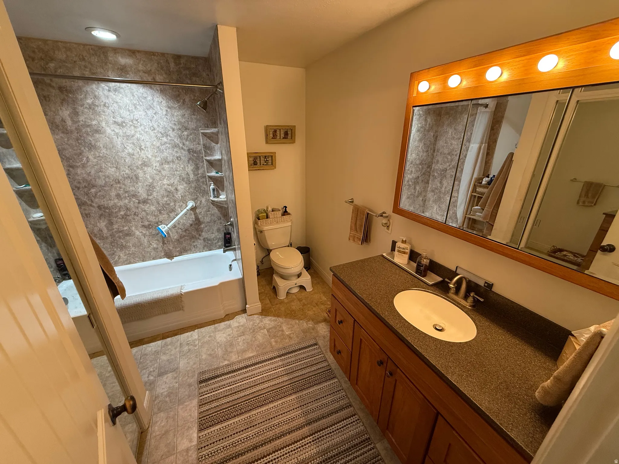 Full bathroom featuring a white tub, textured shower surround with built-in shelving, wood vanity with an integrated sink, dark countertop, and a wood-framed mirror with integrated lighting