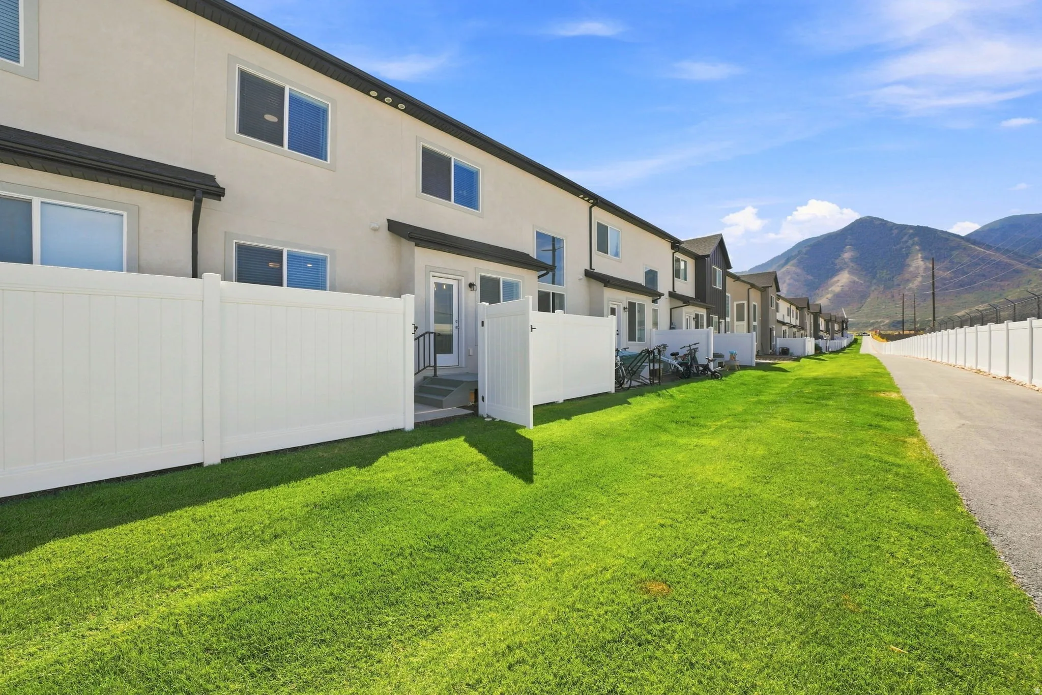 Modern townhome community featuring manicured green space, white privacy fencing, contemporary multi-story architecture with large windows, and mountain backdrop scenery