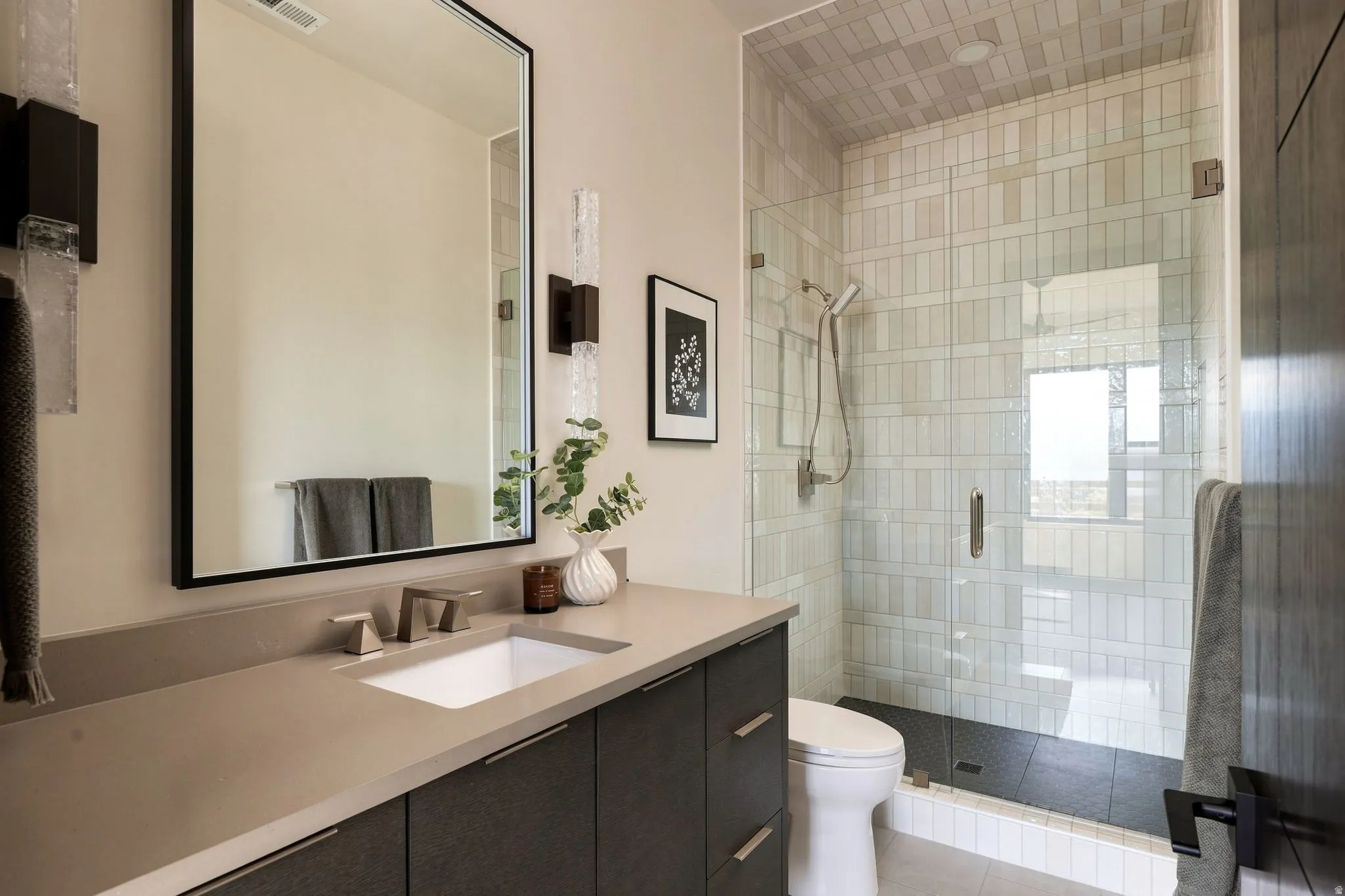 Integrated vanity with a rectangular sink, wall-mounted light fixtures, framed mirror, and a glass-enclosed shower with vertical tile