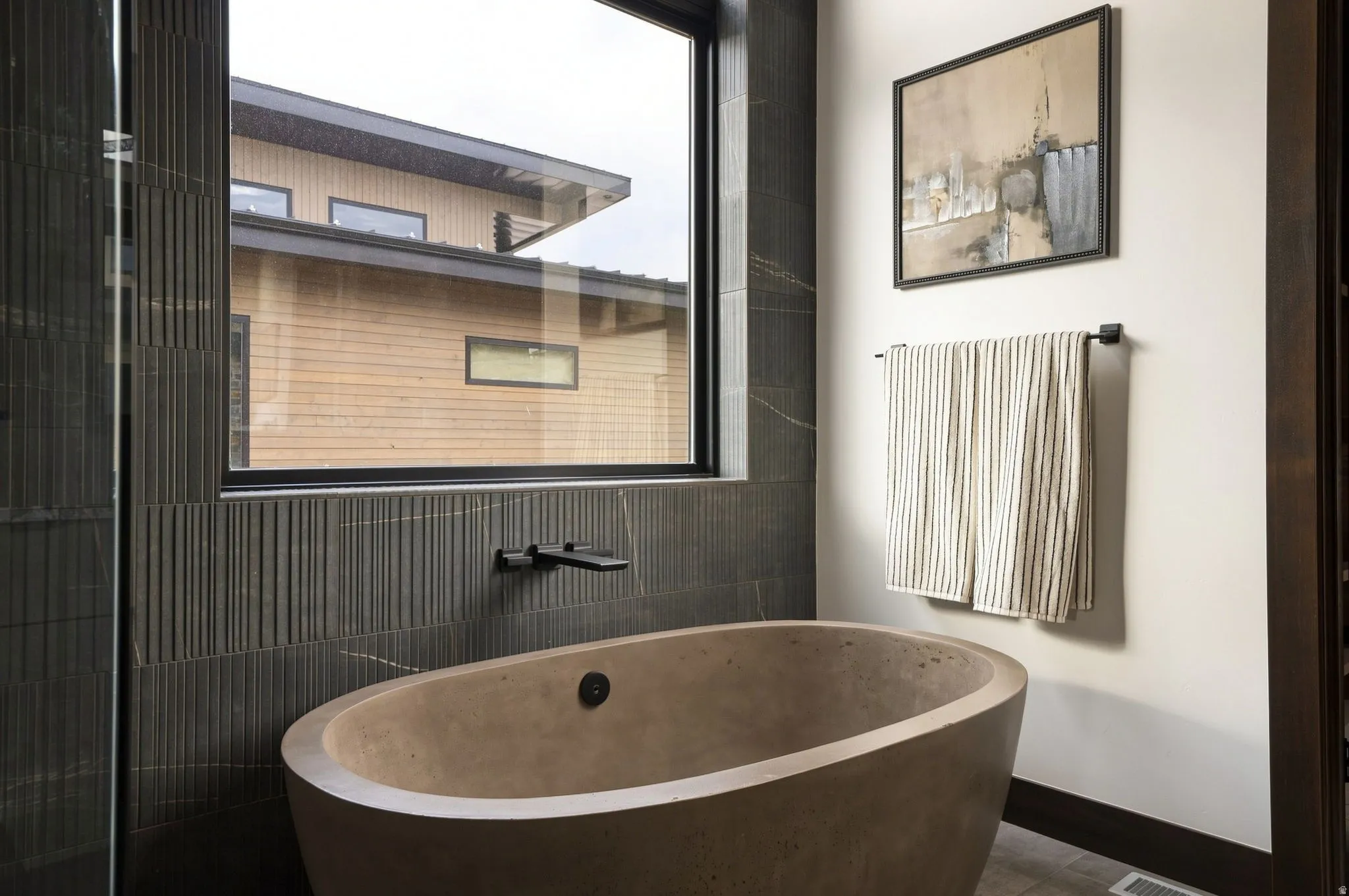 Freestanding soaking tub with dark finish wall-mounted faucet, full-height tile accent wall, and large picture window