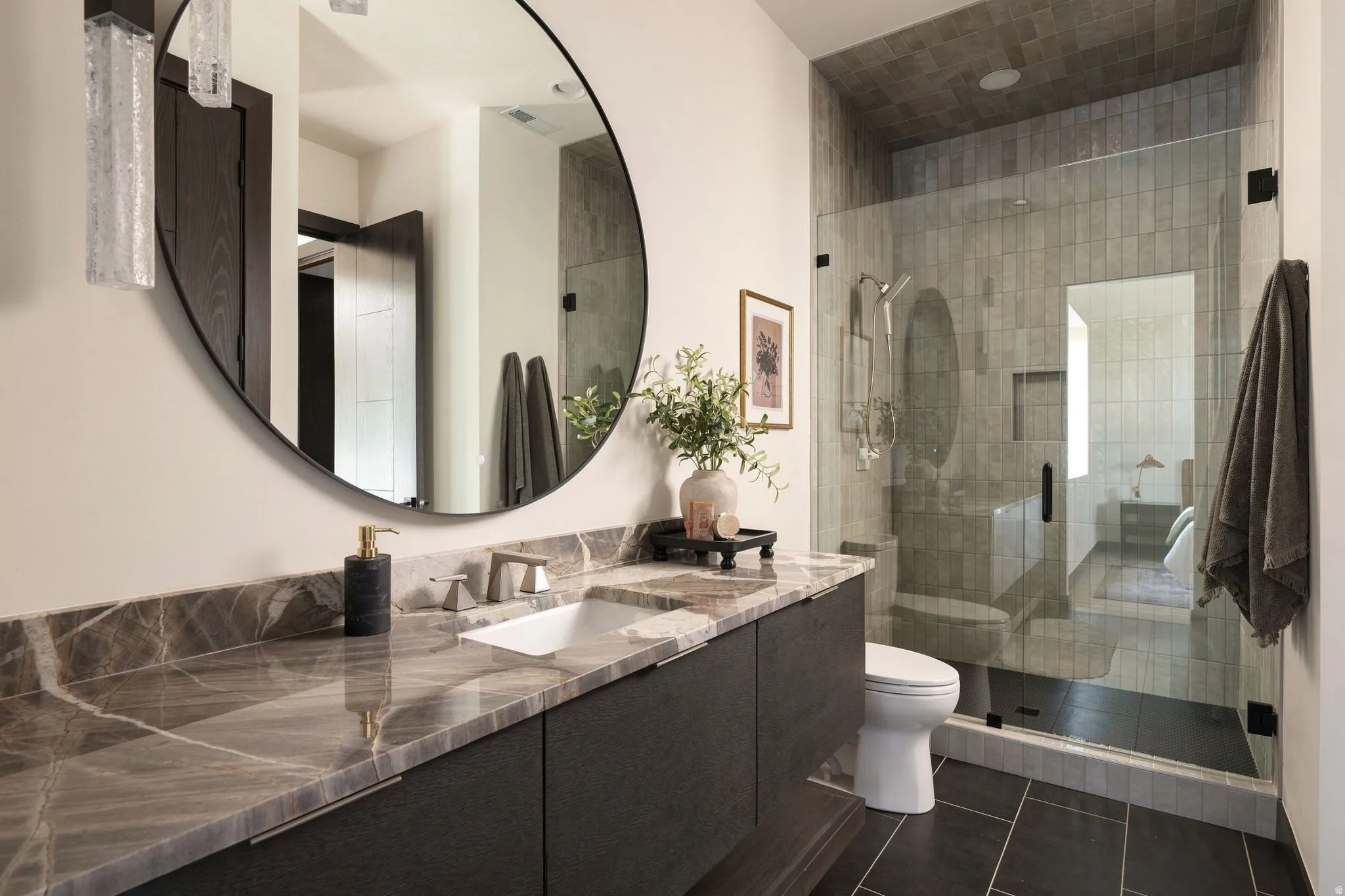 Dual vanity with stone countertop, large round mirror, glass-enclosed shower, rectangular floor tiles, and recessed lighting