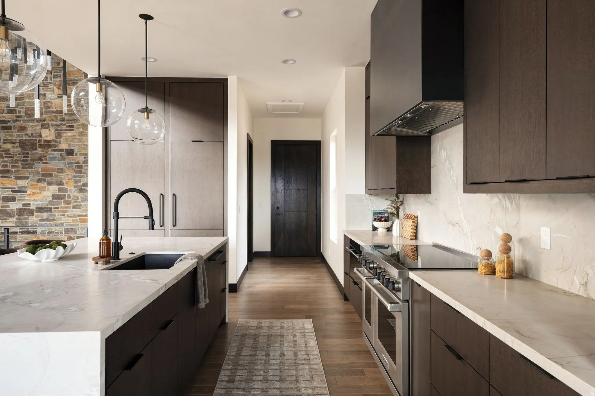Modern kitchen featuring dark wood-finish cabinetry, light stone countertops, stainless steel appliances, and a stone accent wall