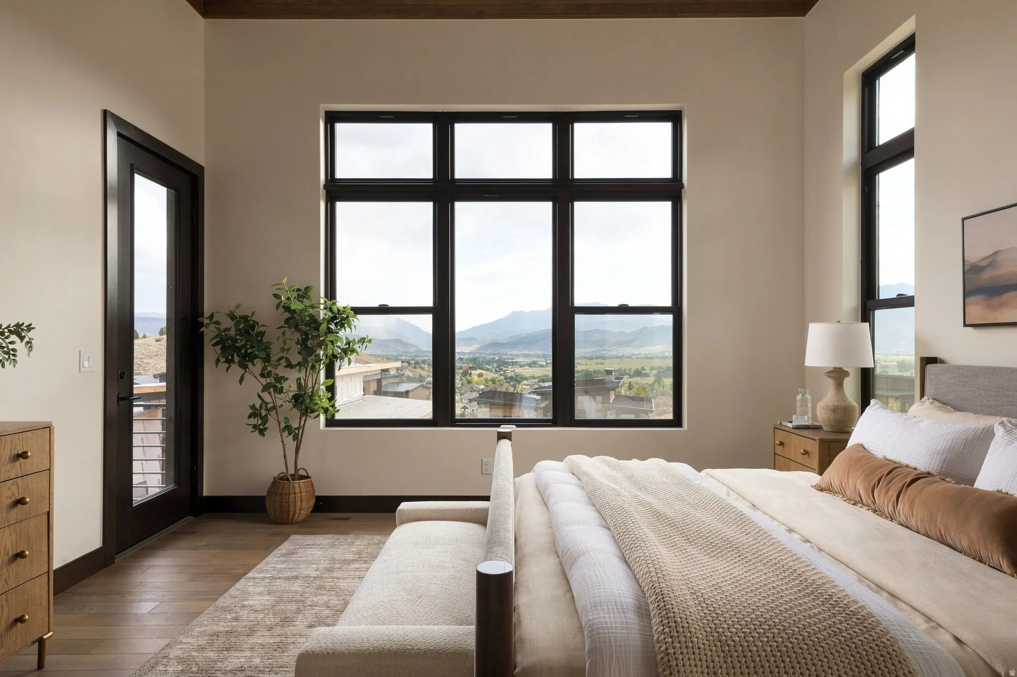 Expansive window wall, wood-finish flooring, dark-trimmed glass door, dark wood ceiling beam, and light-toned walls