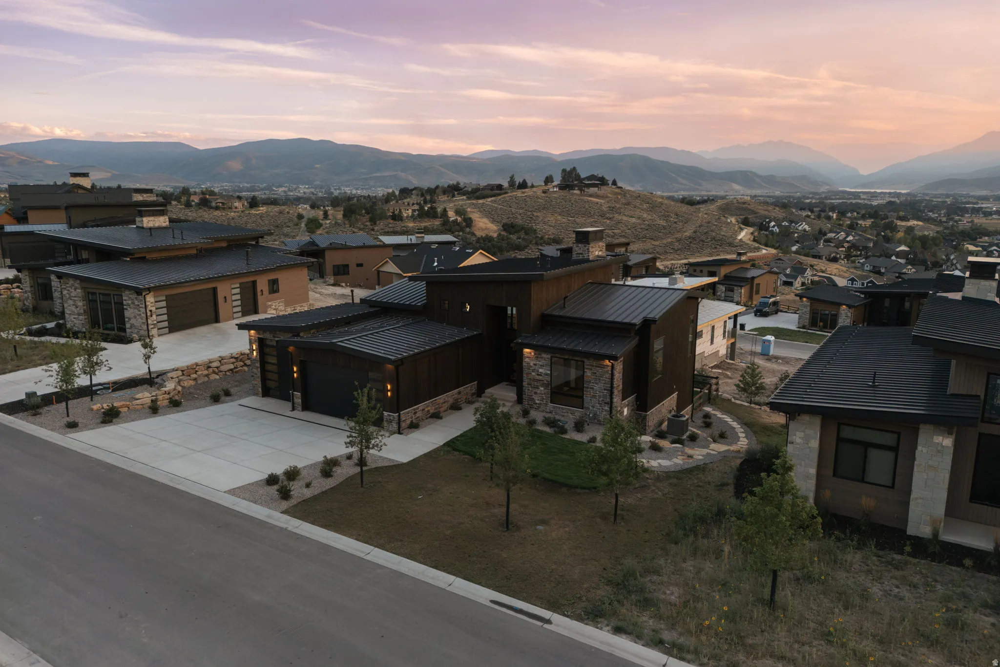 Contemporary architectural style with stone and wood paneling, black standing seam metal roof, paved driveway, and integrated two-car garage