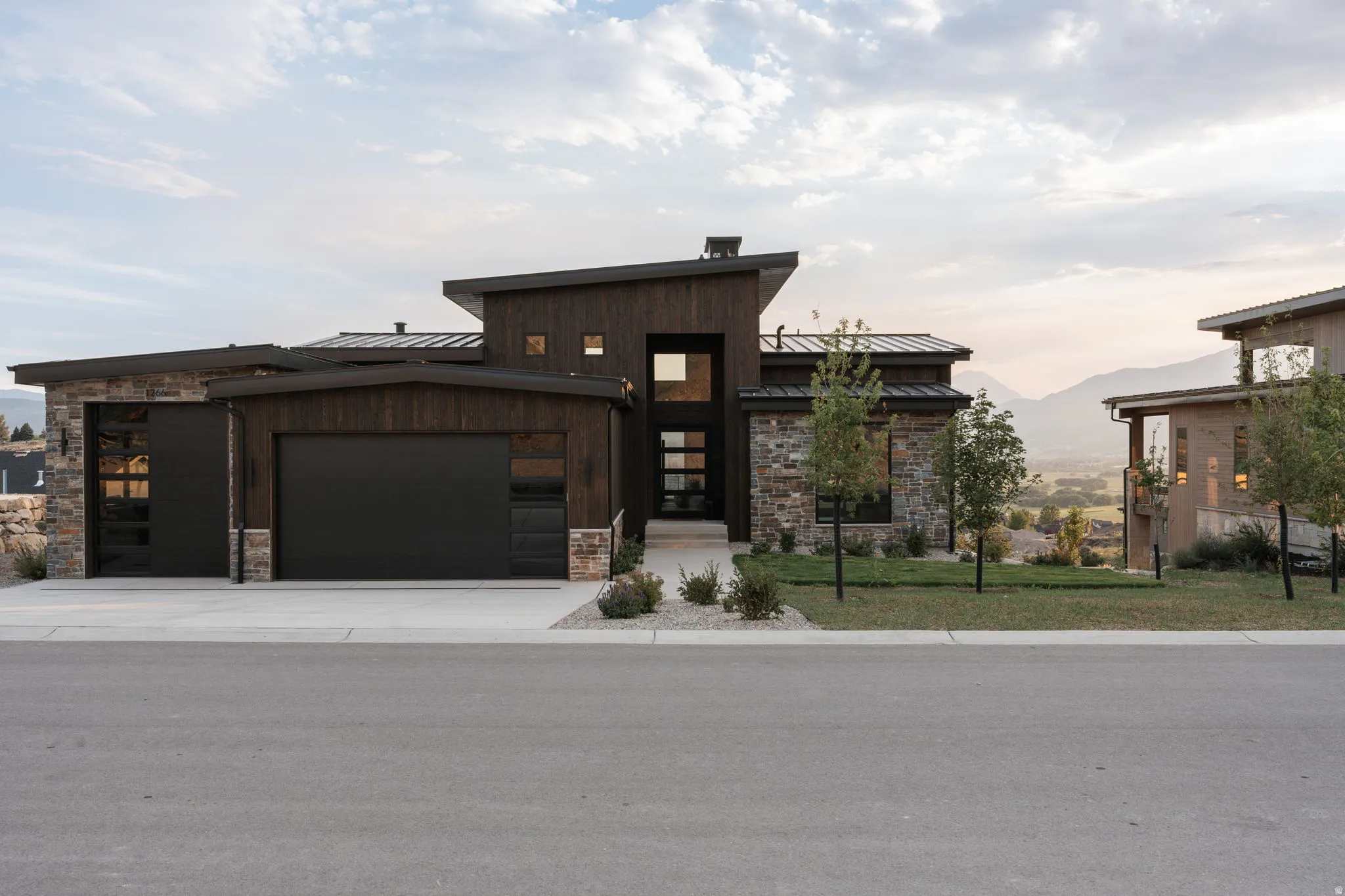 Modern architectural design featuring dark wood-finish siding, natural stone accents, a multi-paneled glass entry door, and dark garage doors with integrated window panels