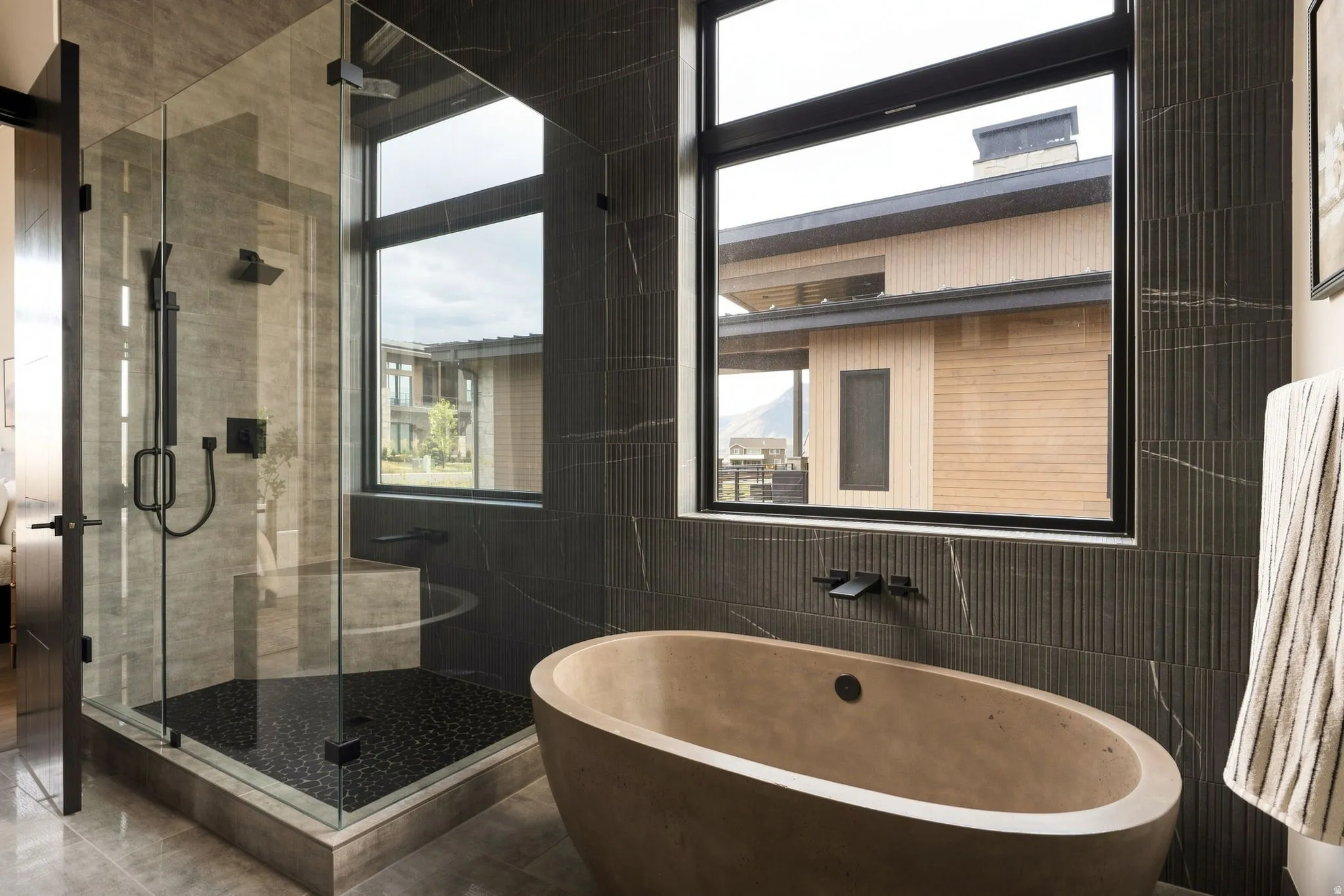 Freestanding soaking tub, glass-enclosed shower with black-finish fixtures, and dark stone-look wall tiling