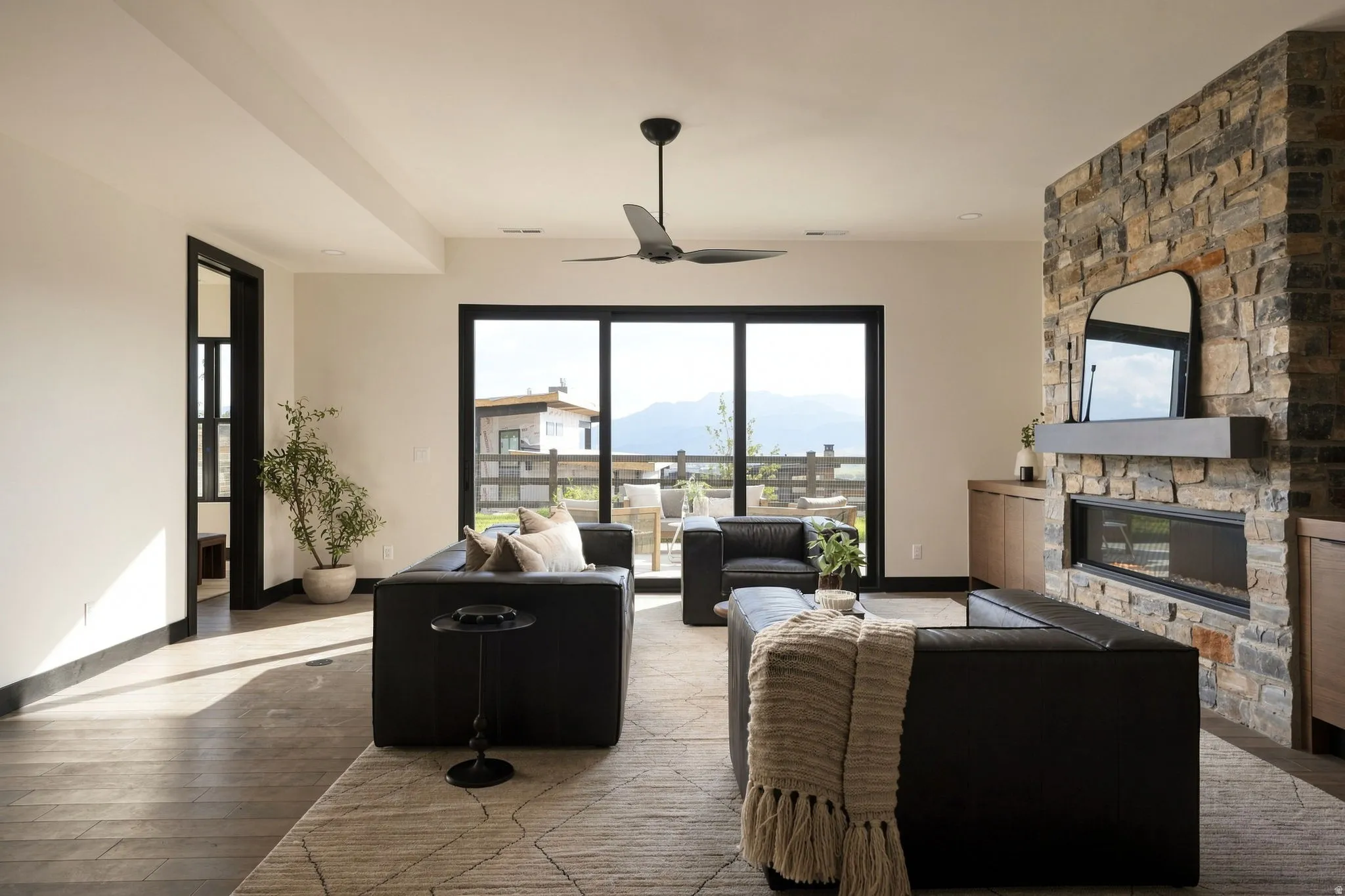Expansive glass sliding doors, stone fireplace with built-in linear electric insert, modern ceiling fan, wood-finish flooring, and black trim throughout