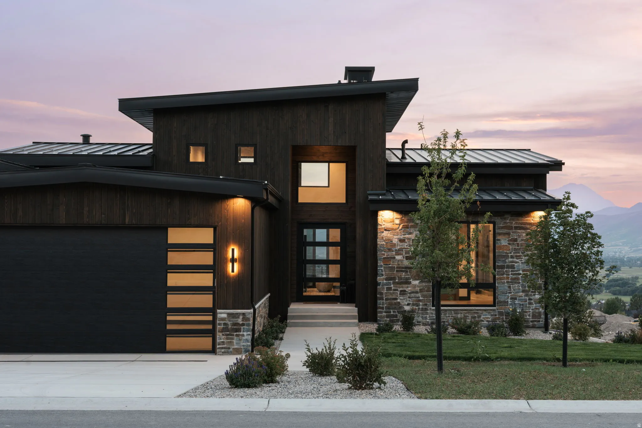 Modern exterior featuring dark wood siding, stone accents, a multi-pane front door, a garage with horizontal window inserts, and a low-sloped roofline