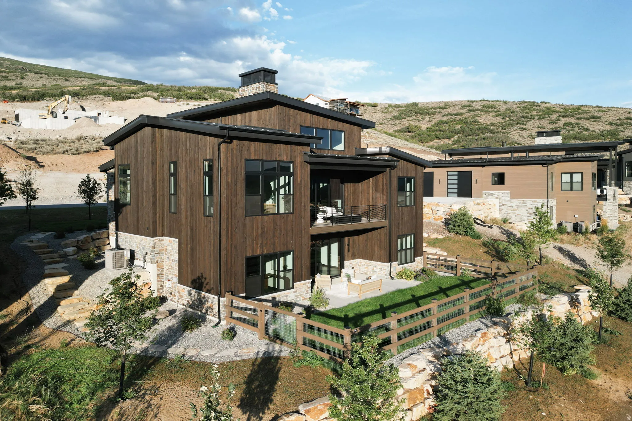 Modern exterior featuring dark wood siding, stone accents, multiple large windows, a ground-level patio, and an upper-level balcony