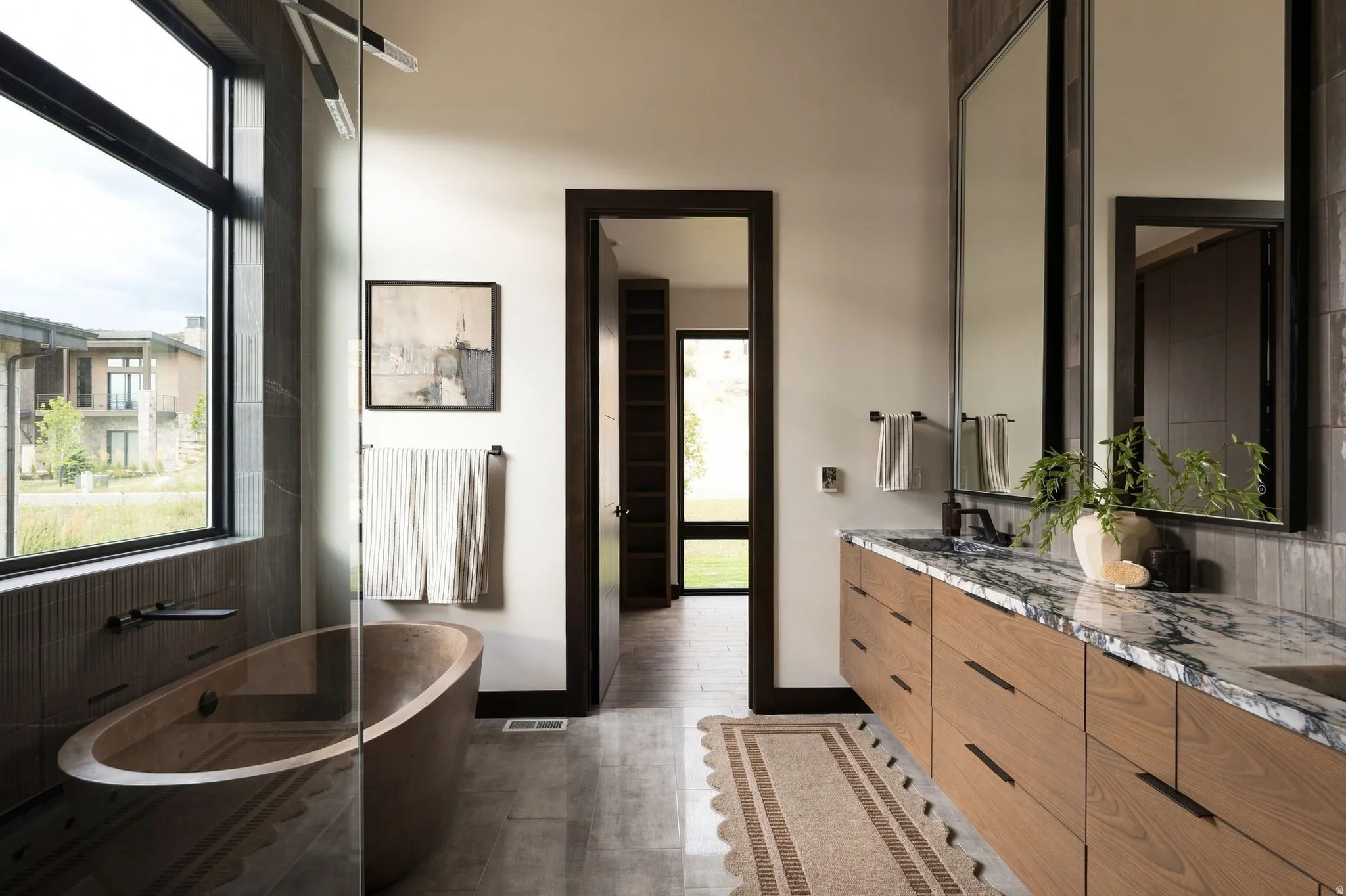 Freestanding soaking tub with dark-framed window, dual vanity with undermount sinks, and stone countertop