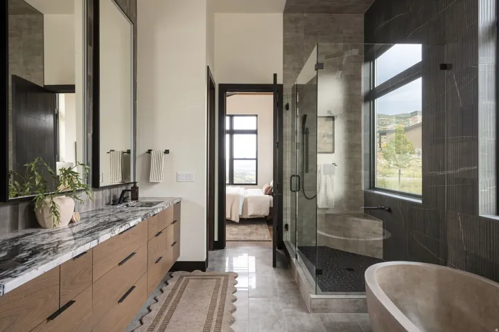 Dual vanity with a stone countertop, built-in soaking tub, glass-enclosed shower, and large windows