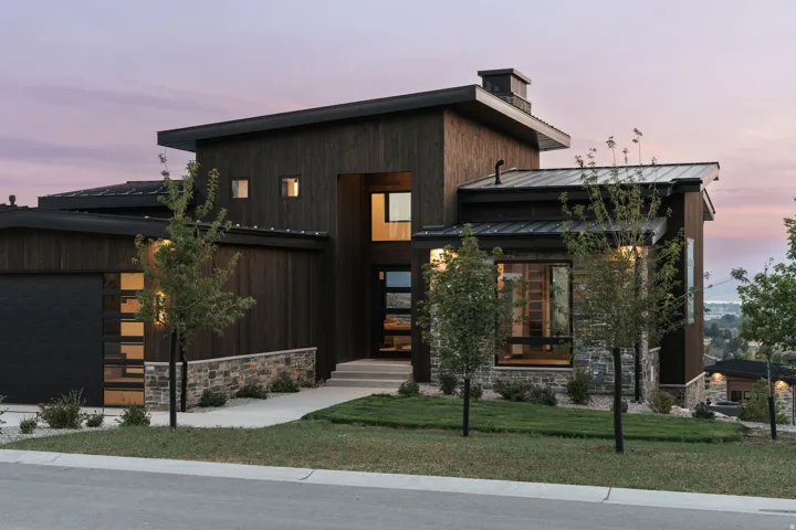 Modern architectural exterior featuring a dark wood-finish facade, natural stone accents, and a sloped metal roof