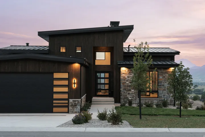 Modern exterior featuring dark wood siding, stone accents, a multi-pane front door, a garage with horizontal window inserts, and a low-sloped roofline