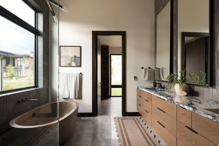 Freestanding soaking tub with dark-framed window, dual vanity with undermount sinks, and stone countertop