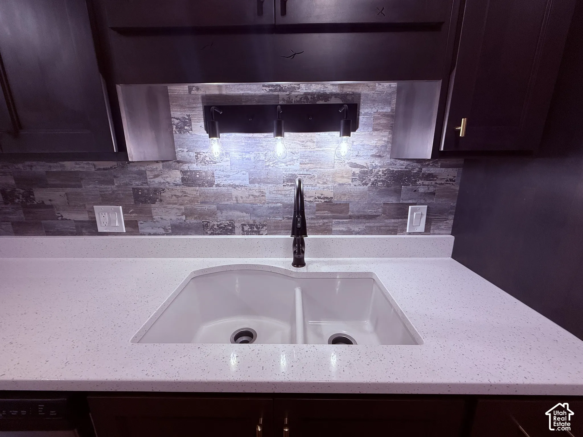 White double-basin sink set into a speckled white countertop, accented by a dark faucet and a stone-look tile backsplash