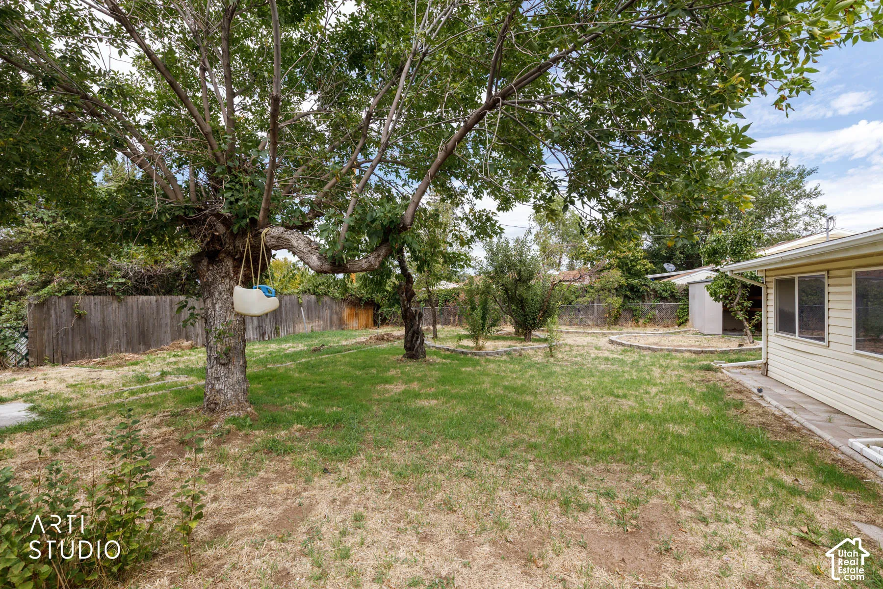 Spacious backyard with mature shade trees and established lawn, anchored by a single-story residence with modern siding and covered porch access